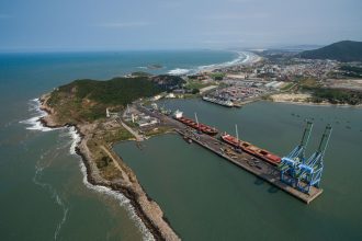 Vista aérea do porto de Imbituba, com navios, cais, guindastes e a faixa litorânea