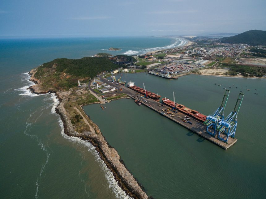 Vista aérea do porto de Imbituba, com navios, cais, guindastes e a faixa litorânea