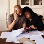 Casal sentado à mesa analisa documentos financeiros enquanto consulta um smartphone.