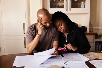 Casal sentado à mesa analisa documentos financeiros enquanto consulta um smartphone.