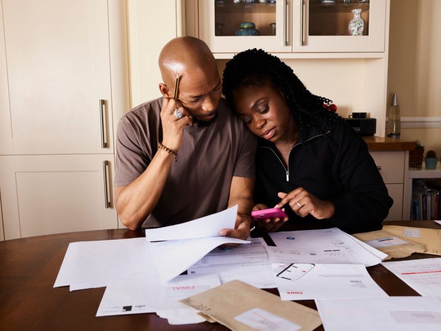 Casal sentado à mesa analisa documentos financeiros enquanto consulta um smartphone.