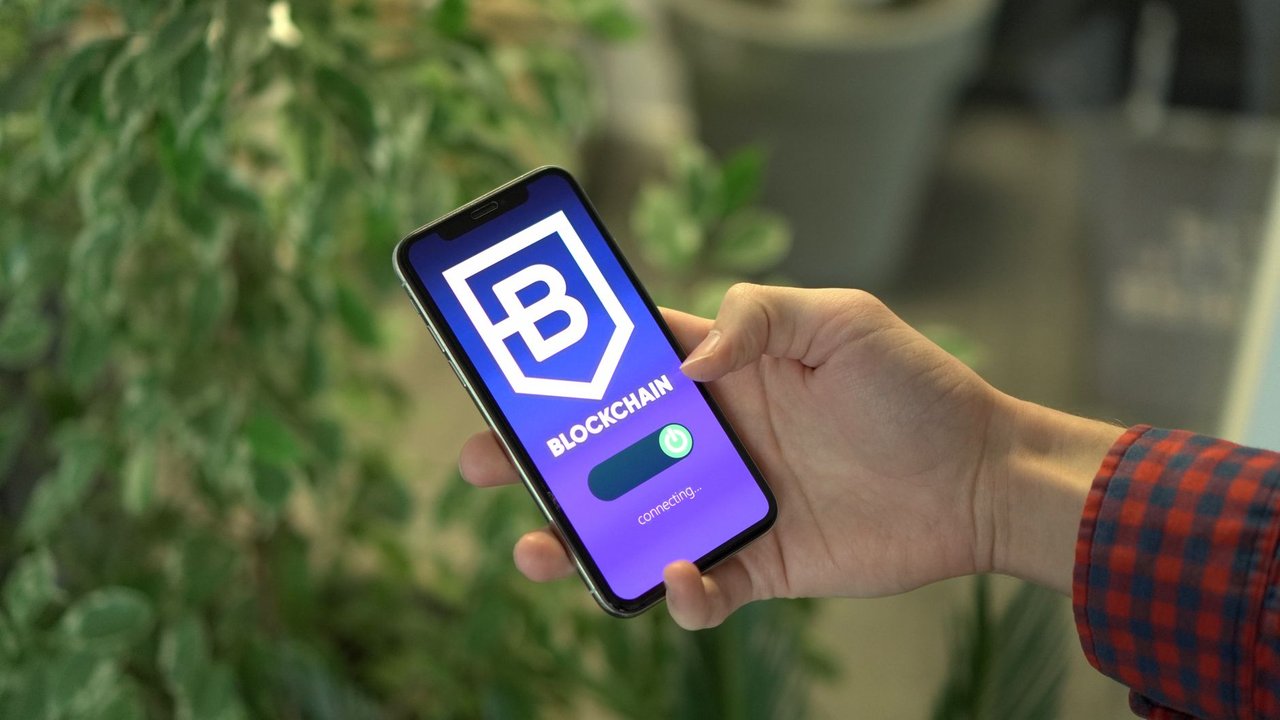 Mão segura um smartphone que exibe a tela de um aplicativo de blockchain.