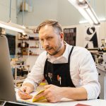 Empresário trabalha diante do computador em uma gráfica movimentada.