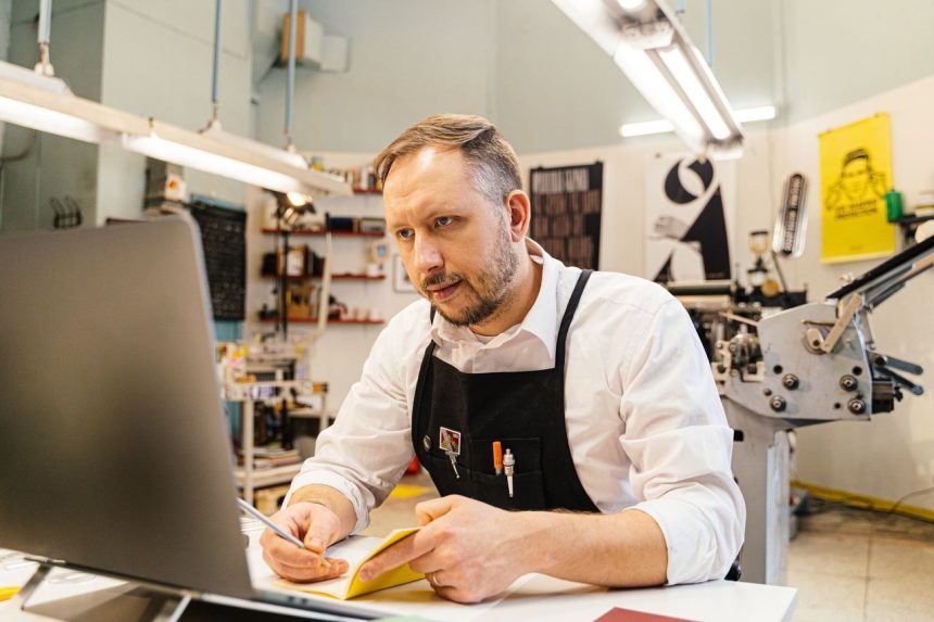 Empresário trabalha diante do computador em uma gráfica movimentada.