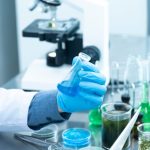 Cientista usando luvas analisa uma amostra azul em laboratório, ao lado de um microscópio e vidrarias.