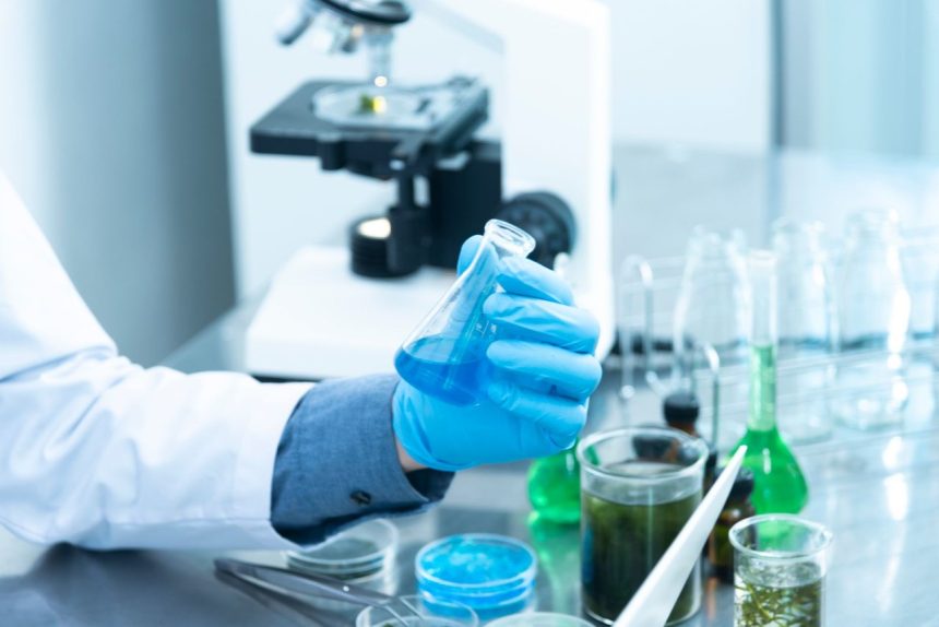 Cientista usando luvas analisa uma amostra azul em laboratório, ao lado de um microscópio e vidrarias.